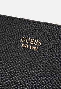 Lompakko mustalla tekstuurilla, jossa on kultainen "GUESS EST 1981" -logo. Varustettu pehmeällä pinnalla ja näkyvillä saumoilla reunoilla.