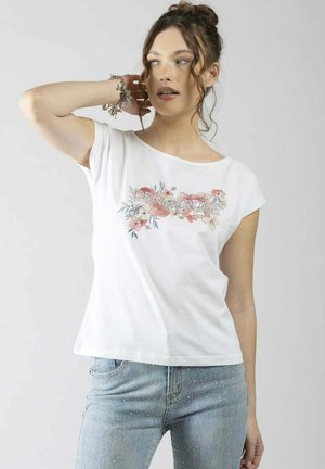 Giovane donna con capelli ricci raccolti in uno chignon indossa una maglietta bianca con motivo floreale e jeans azzurri, posando con la mano vicino alla testa.