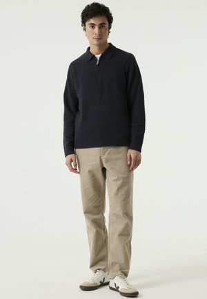 Man standing in neutral pose, wearing a dark zip-collar sweater, beige pants, and white sneakers with black detail against a plain light background.