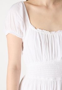 Vit klänning med korta, rynkade ärmar och fyrkantig halsringning. Livet har smockning för textur och framhäver midjan.