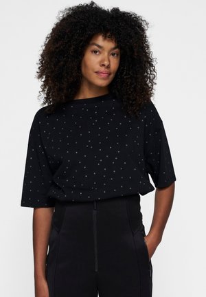Czarna koszula z krótkim rękawem w kropki. Wykonana z miękkiego materiału, o luźnym kroju i dekolcie w łódkę, zestawiona z czarnymi spodniami.
