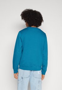 Türkiser Fleece-Pullover mit langen Ärmeln, Rundhals und gerippten Bündchen. Getragen mit hellblauen Cargo-Hosen, die seitliche Taschen haben.