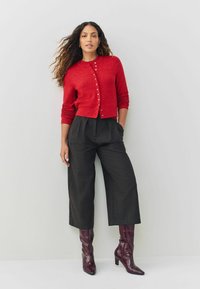 Red knitted cardigan with button front, paired with high-waisted dark gray trousers. Model wears burgundy heeled boots. Smooth texture.