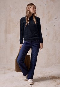 Maglione in tessuto a coste blu scuro, abbinato a jeans blu. Porta una borsa imbottita marrone. Sneakers con accenti bianchi e beige. Sfondo semplice.