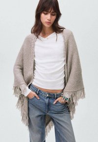 Light gray knitted shawl with fringe edges, worn over a white long-sleeve top, paired with light blue denim jeans.