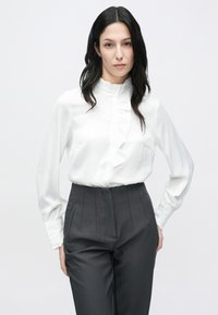 Balta blūze ar garām piedurknēm, dekorēta ar volāniem un ar pogu aizdari, kopā ar tumši pelēkām biksēm ar augstu jostasvietu, kas ir ar vertikālām šuvēm un kabatām.