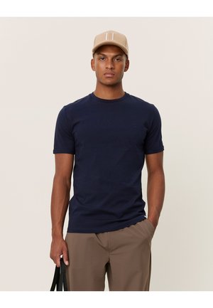 T-shirt a maniche corte blu navy realizzato in cotone, con scollo a girocollo e piccolo logo decorativo. Indossato con berretto beige e pantaloni color cammello.
