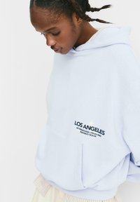 Felpa oversize azzurro chiaro con tasca frontale, caratterizzata da un testo nero e oro: "LOS ANGELES" e un emblema. Realizzata in tessuto morbido.