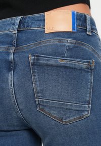 Mørkeblå denimjeans med en enkel baklomme og et brunt lærmerke med blå detaljer sydd på midjebåndet.