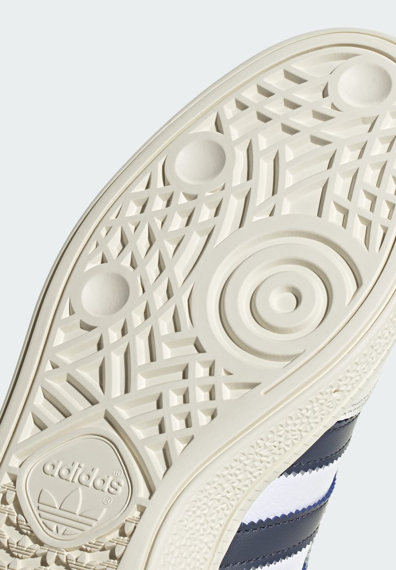 Podeszwa sneakersów Adidas z teksturowanej białej gumy z okrągłymi i geometrycznymi wzorami, logo oraz gładkimi krawędziami.