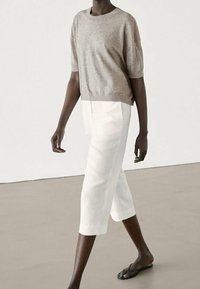 Vaaleanruskea lyhythihainen neule yhdistettynä valkoisiin lyhyisiin housuihin ja mustiin flip-flop-sandaaleihin, esittäen rentoa istuvuutta ja sileää materiaalia.