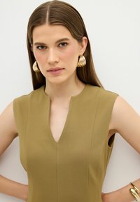 Olivengrünes, ärmelloses Kleid mit V-Ausschnitt und taillierter Passform, kombiniert mit großen goldenen Creolen. Glatte Stoffstruktur.