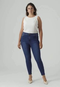 Jeans skinny din denim albastru cu talie înaltă, asortați cu un top alb cu nervuri fără mâneci și tocuri bej, stând pe un fundal neutru.