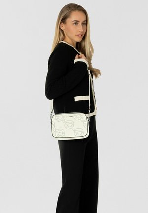 Bolso bandolera en blanco con patrón delineado en negro. Presenta una forma rectangular, material sintético suave y correa ajustable.
