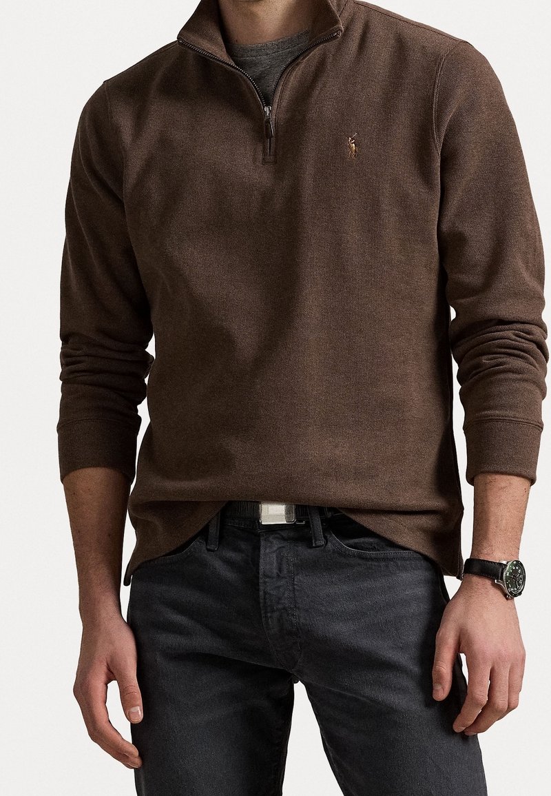 Homme portant un pull marron à demi-fermeture éclair, un jean foncé avec ceinture, et une montre-bracelet, debout devant un fond clair uni.