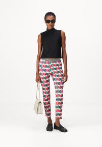 Schwarzes ärmelloses Oberteil, bedruckte schlanke Hose im geometrischen Muster in Pink, Grün und Weiß, und schwarze Loafer; hält eine leichte Handtasche.