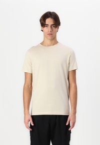 Bežs īsām piedurknēm t-krekls, izgatavots no mīksta auduma ar apaļu izgriezumu, kuru valkā modelis uz vienkrāsaina baltā fona, kombinēts ar melnām biksēm.