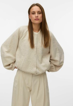 Jeune femme aux longs cheveux bruns portant une veste matelassée crème à manches ballon et un pantalon beige taille haute, debout les mains dans les poches.