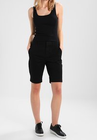 Débardeur noir associé à un short noir ; le short comporte des poches latérales et une coupe ajustée. Porté avec des baskets noires. Design simple et minimaliste.