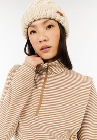 Bruine en crème gestreepte kwart-zip top met een hoge kraag, gemaakt van een structuurstof. Model draagt een crème gebreide beanie met een leren label.