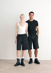 Shorts noirs avec cordon de serrage en coupe décontractée, portés par deux modèles. L'un porte un débardeur blanc à côtes et l'autre un t-shirt noir, tous deux associés à des bottes.