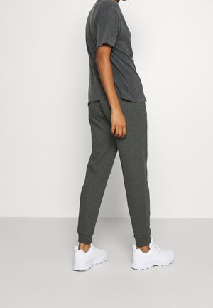 Personne portant un pantalon de jogging gris foncé, un t-shirt gris foncé assorti et des chaussures de sport blanches, debout contre un mur blanc uni.