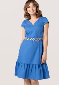 Blauwe katoenen jurk met een V-hals, korte mouwen en een gerimpelde onderkant. Versterkt met een ronde metalen riem rond de taille.