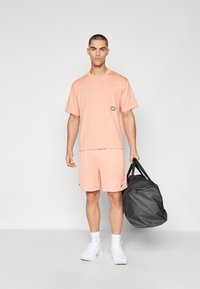 Tricou sportiv roz cu mânecă scurtă și pantaloni scurți, croială lejeră. Modelul ține o geantă de sport neagră, purtând adidași și sosete albe.