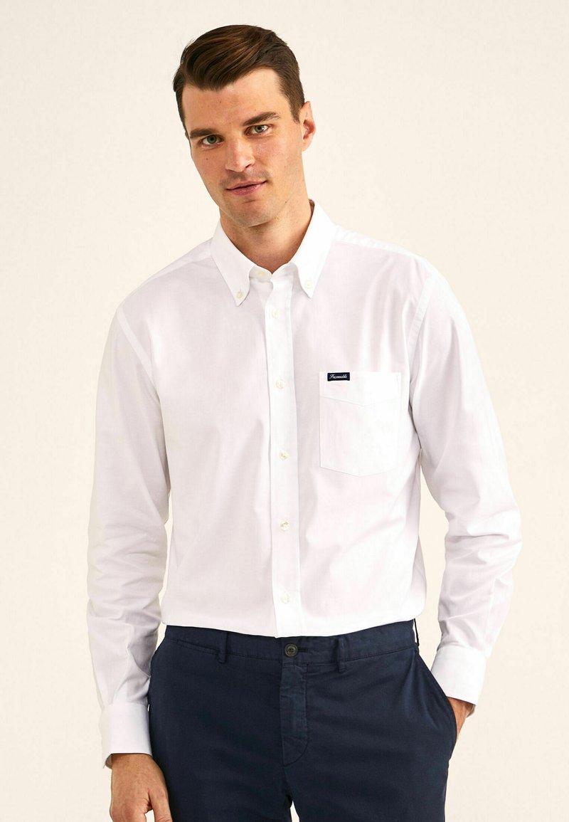 Camisa blanca de botones con un solo bolsillo en el pecho y una pequeña etiqueta de la marca. Mangas largas y cuello, combinada con pantalones azul marino oscuros.