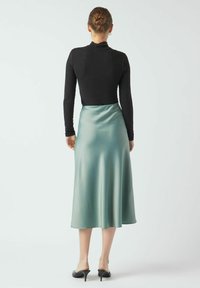 Must have pikad varrukad ja must topp, mis on kombineeritud rohelise satiinist midi-seelikuga. Seelikul on sile tekstuur ja see ulatub kesk sääreni, veidi kogutud taljejoonega.