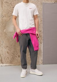 Vit Nike t-shirt med liten logotyp, rosa sweatshirt knuten runt midjan, grå byxor och vita sneakers med grå accenter. Texturerad vägg i bakgrunden.