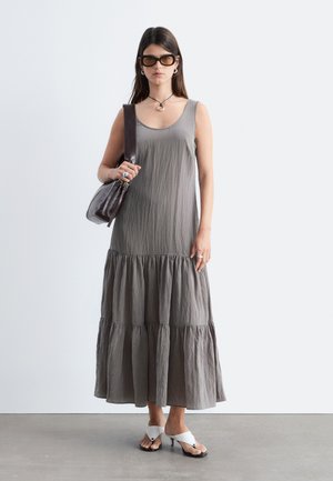 Femme portant une robe maxi grise sans manches à volants, des sandales blanches à talons, de grandes lunettes de soleil, des boucles d'oreilles créoles, une bague, un collier et portant un sac à main marron.