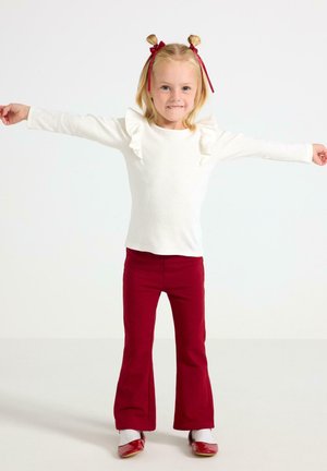 Valkoinen pitkähihainen paita, jossa on röyhelöt olkapäillä, yhdistettynä punaisiin leveisiin housuihin ja punaisiin kenkiin. Lapsi seisoo käsivarret ojennettuna, hymyillen.