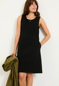 Schwarzes ärmelloses Kleid mit figurschmeichelnder Silhouette, seitlichen Taschen und rundem Ausschnitt. Das Model hält eine olivgrüne Wildlederjacke.