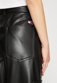 Mustat nahkahousut, joissa on takatasku, jossa on erottuva Tommy Jeans -logopatchi. Housuissa on sileä rakenne ja säädetty muotoilu.