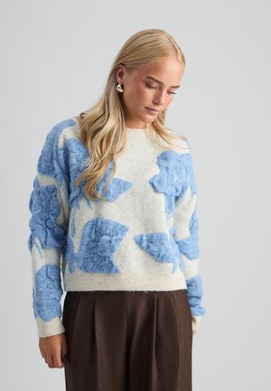 Donna con lunghi capelli biondi che indossa un maglione bianco con grandi fantasie floreali blu texture e pantaloni marrone scuro, guarda verso il basso.