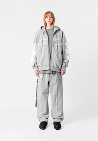 Sudadera gris con cremallera y gráficos blancos en las mangas, combinada con pantalones de chándal grises que tienen un bolsillo lateral. Llevada con zapatos negros texturizados.