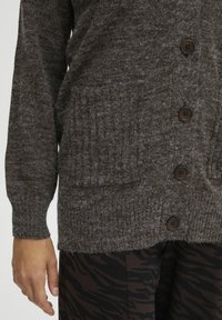 Cardigan a maglia marrone con tessuto strutturato, bottoni frontali e tasche laterali. Abbinato a pantaloni neri con un sottile motivo a righe di tigre.