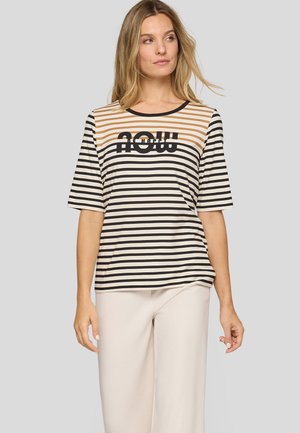 Woman wearing a beige and black striped short-sleeve shirt with the word "NOW" on the chest, paired with light beige pants.