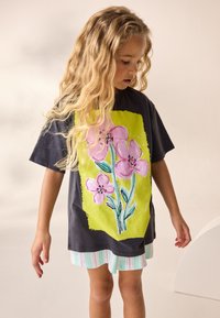 Juoda trumpomis rankovėmis marškinėliai su spalvingu gėlių raštu ant ryškiai žalio fono, derinami su dryžuota šviesios spalvos sijonu.