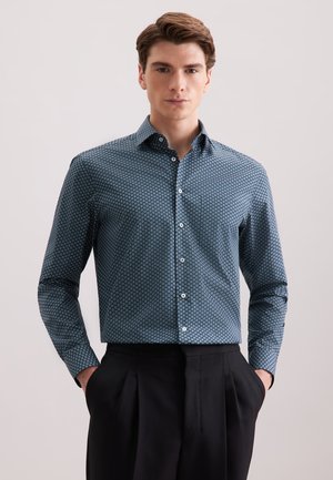 Camicia button-up di colore blu scuro con un motivo geometrico, maniche lunghe e colletto classico. Il tessuto appare liscio. Indossata con pantaloni neri.