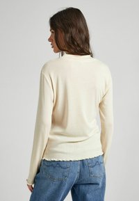 Camicia a maniche lunghe color crema realizzata in tessuto morbido, con scollatura rotonda e orlo scallop, abbinata a jeans in denim blu.