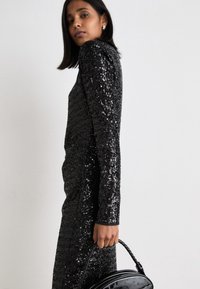 Vestido preto com lantejoulas, mangas longas, silhueta justa e superfície texturizada. Acompanhado por uma mala de mão preta brilhante com formato arredondado.