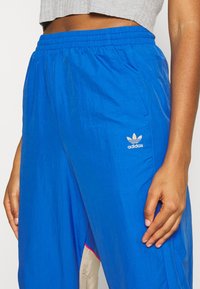 Blå träningsbyxor gjorda av lättviktig material med elastisk midja. Har Adidas-logotypen och kontrasterande beige och rosa detaljer.