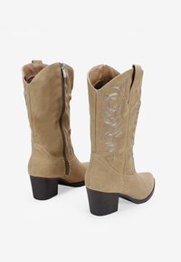 Stivali da cowboy in suede beige con punta affusolata, tacco medio e ricami decorativi in argento sulla gamba. Cerniera laterale per una facile vestibilità.
