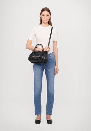 Bolso de mano negro acolchado con una placa de logo dorada, que presenta dobles asas y una correa de hombro desmontable. Combinado con jeans de color azul claro y una blusa blanca.