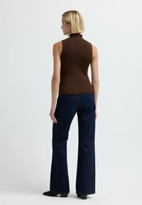 Vrouw met kort blond haar, gekleed in een mouwloze bruine top, donkerblauwe wijde spijkerbroek en bruine hakschoenen, van achteren bekeken tegen een effen achtergrond.
