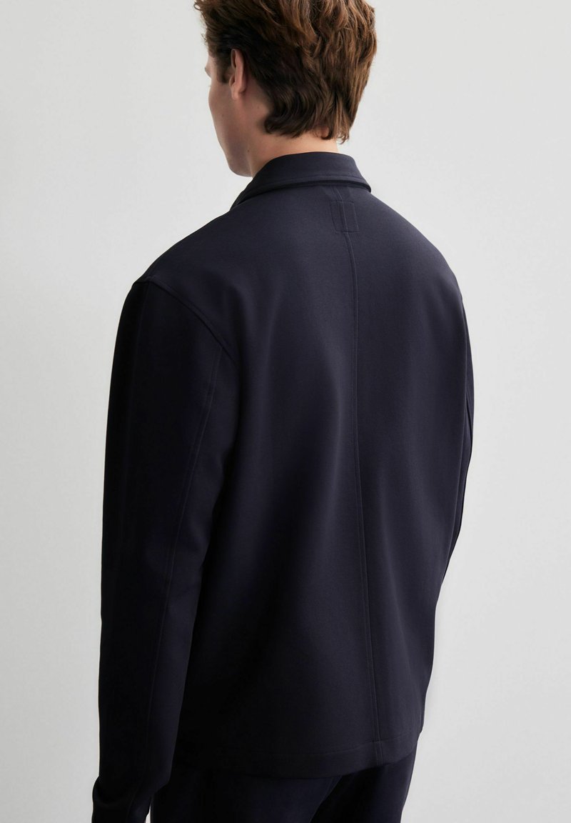 Dunkelblaues Jacket aus glattem, strukturiertem Stoff, mit klassischem Kragen und geradem Schnitt. Sichtbare Nahtdetails auf der Rückseite.
