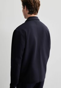 Chaqueta de manga larga azul marino con cuello, textura suave y detalles de costura sutiles, mostrada desde la parte posterior sobre un fondo claro.