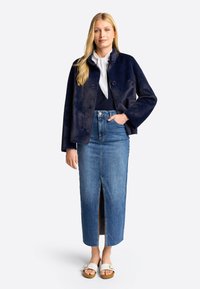 Jachetă din blană ecologică bleumarin cu butoane mari, asortată cu o fustă din denim cu talie înaltă, care are o fante în față și purtată cu sandale albe tip slide.